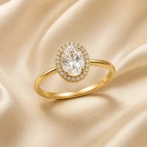 Gold-tone oval halo ring with clear gemstone-style facets on ivory satin